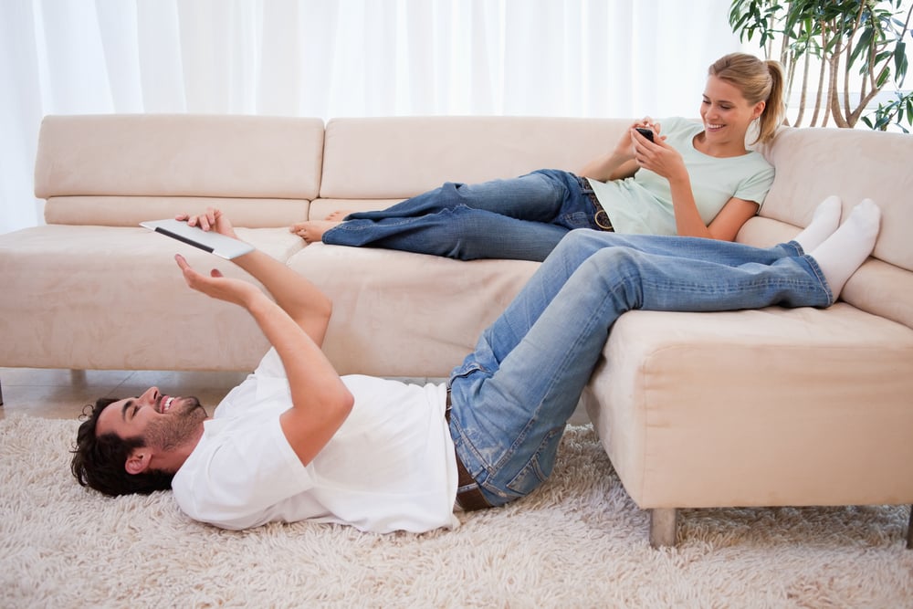 Woman using her smartphone while her boyfriend is using a tablet computer in their living room